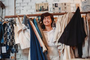 cheerful asian female customer standing among hanging clothes in store and smiling