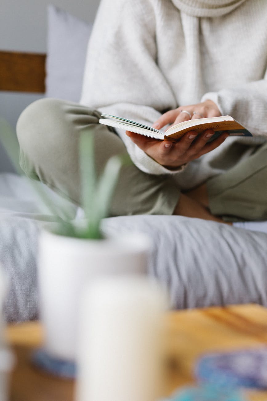 faceless woman reading book on comfortable bed at home