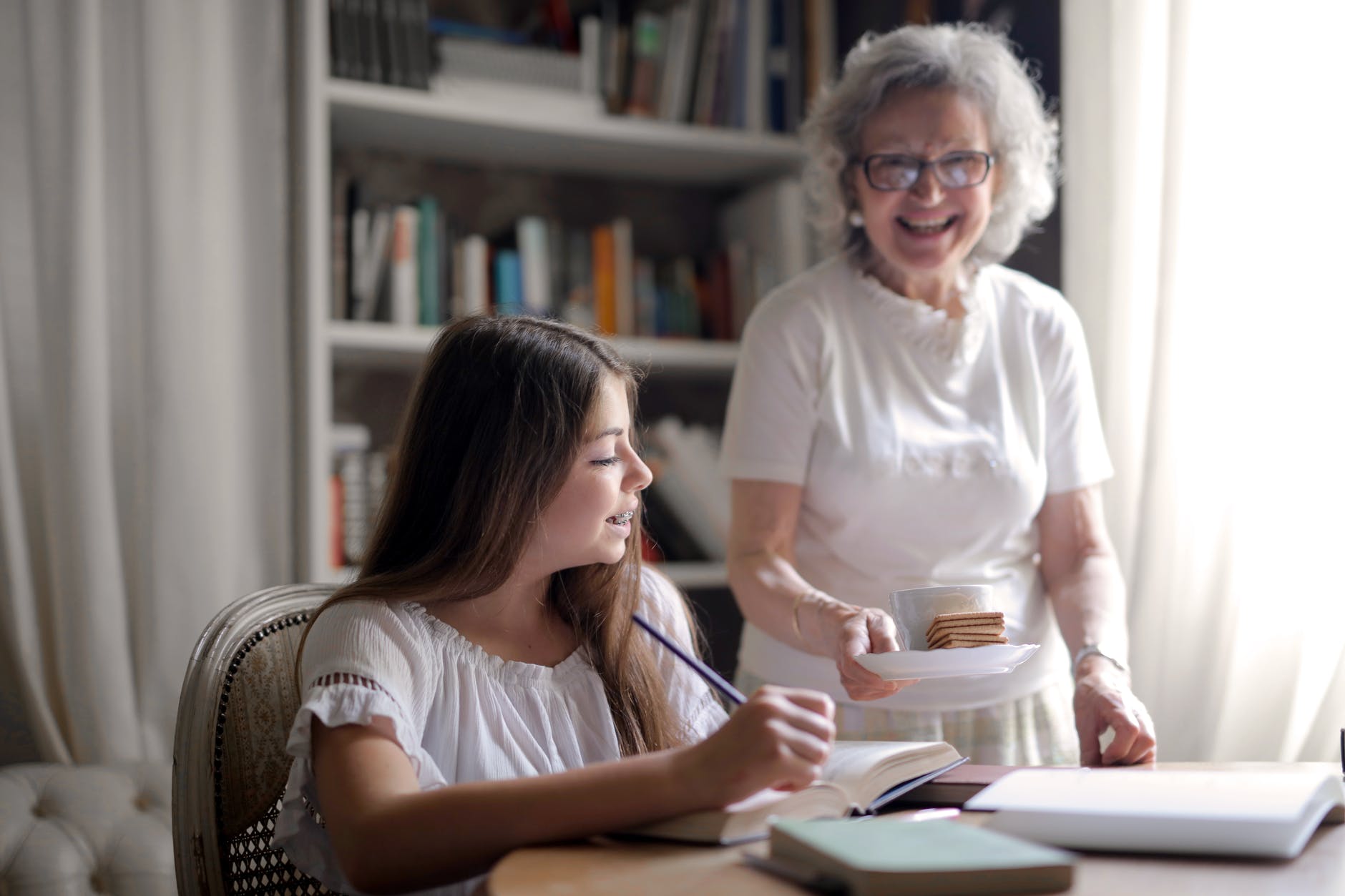 happy aged woman giving cookies and cup of hot drink to pleased granddaughter doing homework at home